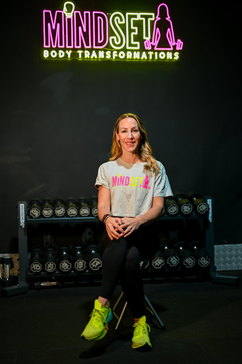 Carrie, founder and head coach of Mindset Body Transformations, seated under the studio neon sign